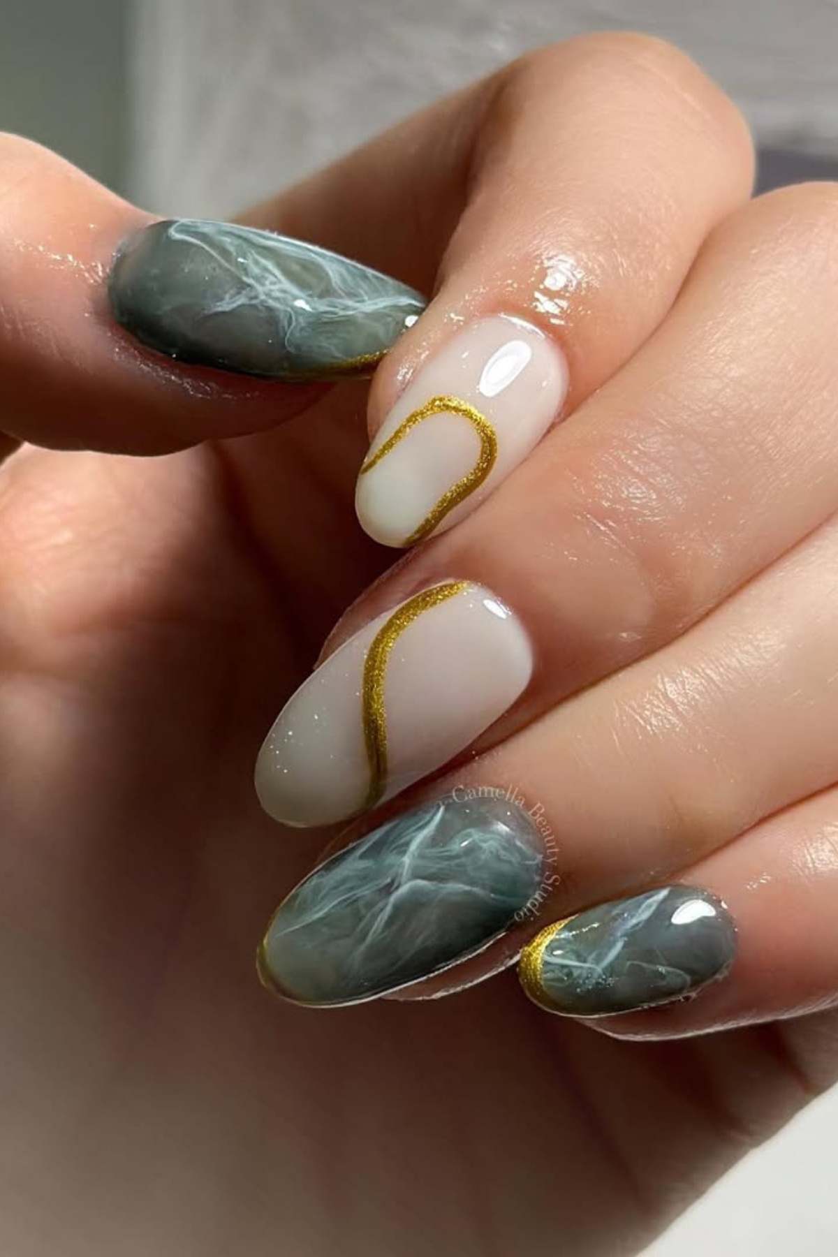 A hand with manicured nails featuring a mix of marble green designs, nude polish, and gold accent lines.