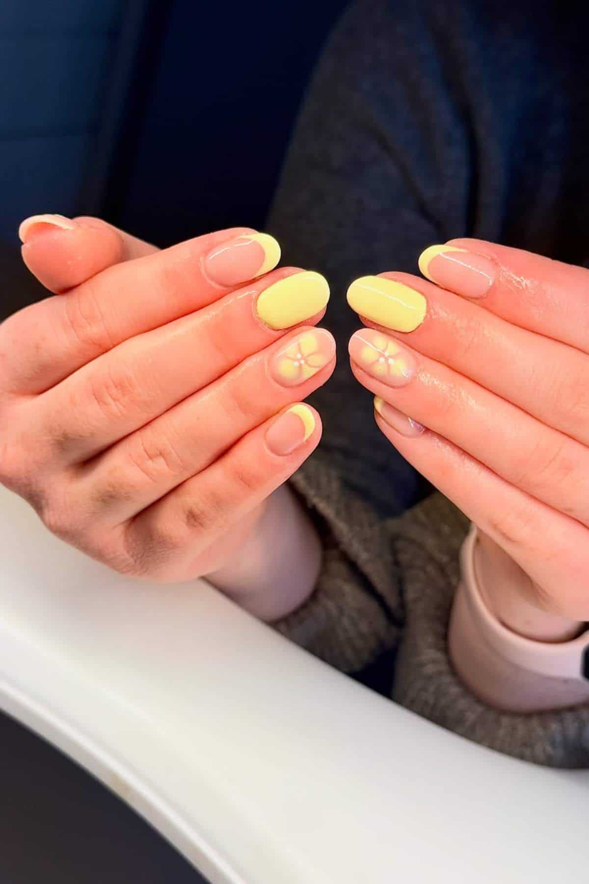 Hands with yellow French tip manicure; ring finger nails feature a simple white flower design, while the rest have plain yellow tips.