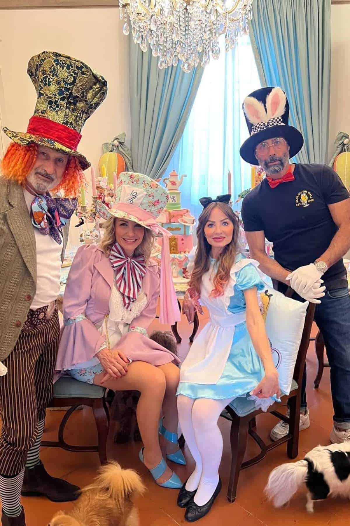 Four adults in Alice in Wonderland costumes pose indoors under a chandelier, with two seated and two standing. Pets are visible at their feet.