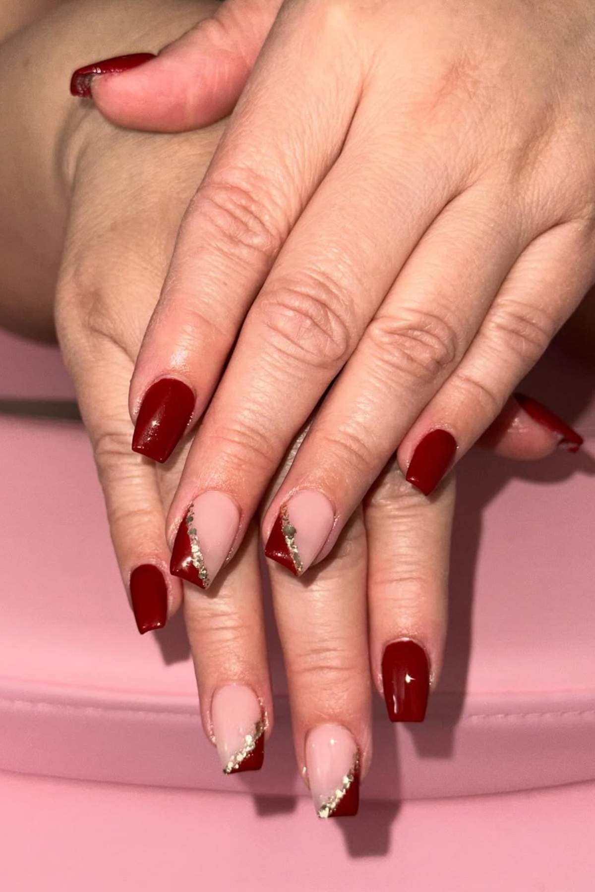 Hands with manicured nails featuring a mix of solid red polish and nude polish with red tips and gold diagonal accents, displayed on a pink surface.