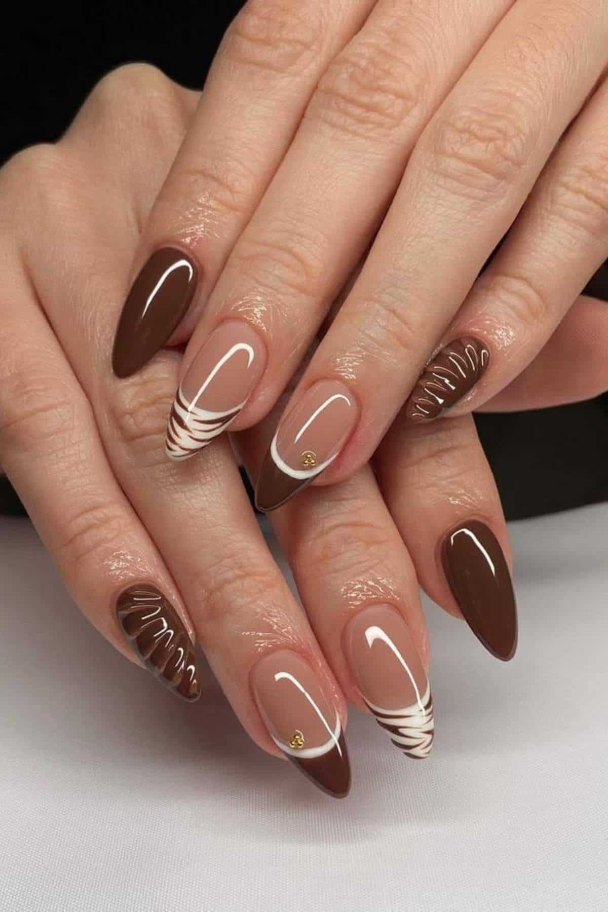 Hands with almond-shaped nails featuring glossy brown polish, some nails with white swirl patterns and small gold studs, displayed on a white surface.