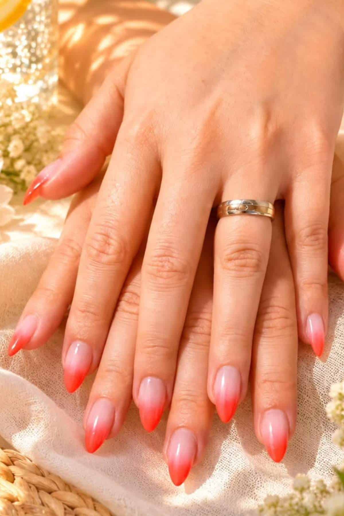 Two hands with long, almond-shaped nails featuring a pink gradient manicure, one hand wearing a silver ring, resting on a light-colored fabric.