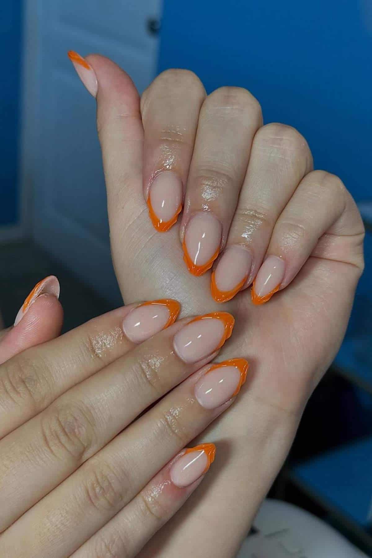 Hands with almond-shaped nails featuring a nude base and bright orange French tips, displayed against a blue background.