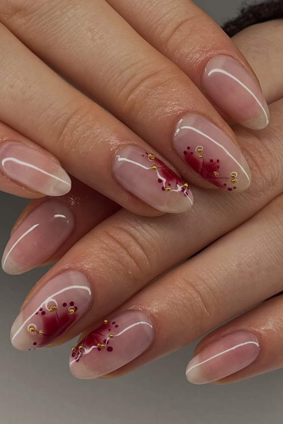 Hands with almond-shaped nails featuring a clear base and floral nail art in red and gold accents on the ring and middle fingers.