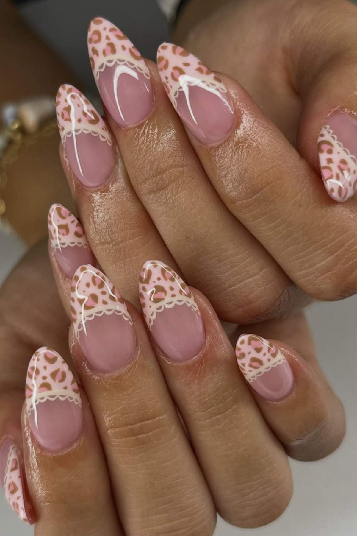 Close-up of hands with almond-shaped nails featuring a pink base, leopard print French tips, and white lace-like accents.