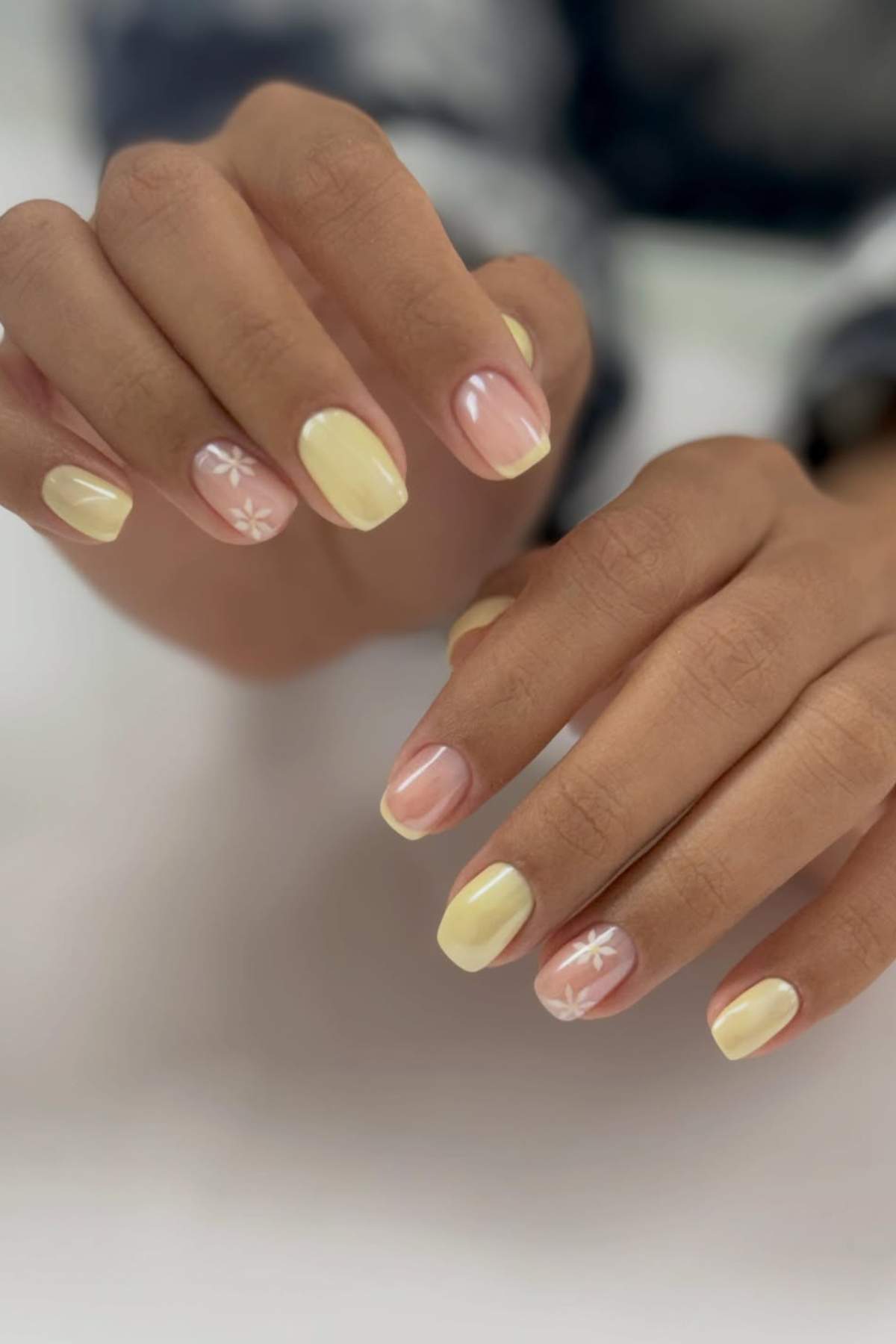 Hands with short, rounded nails painted light yellow, with two accent nails featuring white daisy flower designs on a natural base.