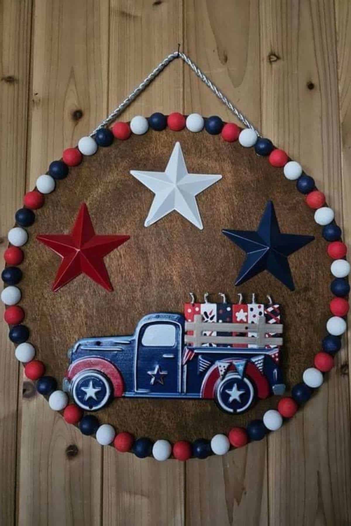A decorative wall hanging features a red, white, and blue truck with stars and gift boxes, surrounded by patriotic beads and three stars on a wooden background.