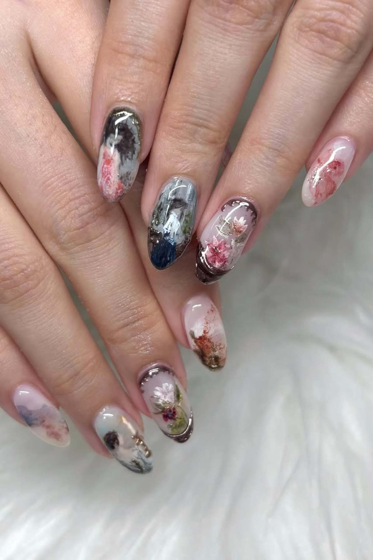 Close-up of hands with almond-shaped nails featuring various abstract floral and marble designs in muted tones on a white faux fur surface.