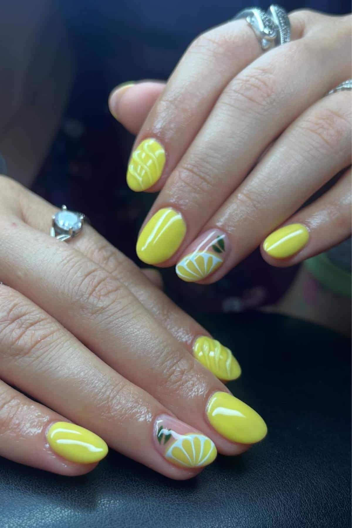 Hands with short, almond-shaped nails painted yellow, featuring lemon and geometric designs on some accent nails. The person is wearing silver rings.