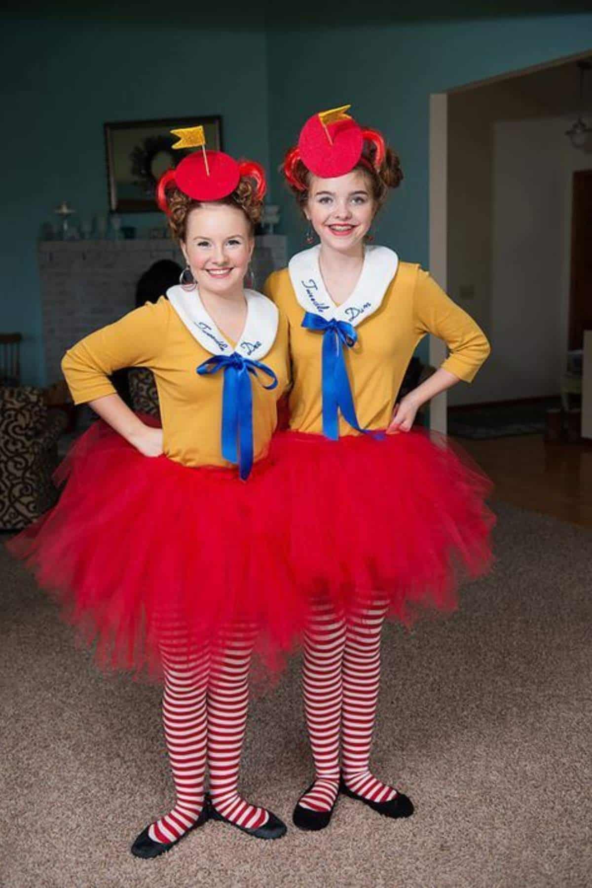 Two people stand indoors wearing matching costumes with yellow tops, blue bows, red tutus, red-and-white striped tights, and red hats resembling Tweedledee and Tweedledum.
