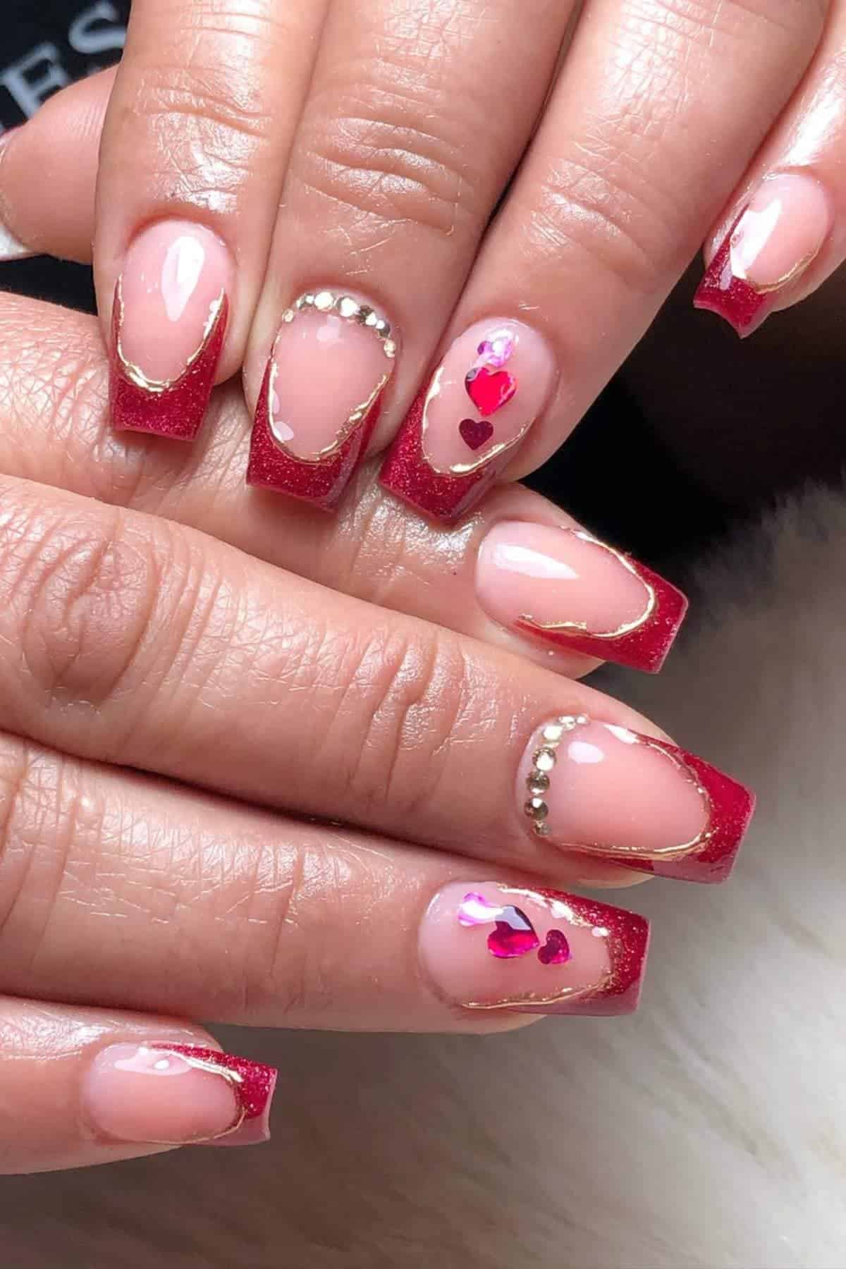 Hands with red glitter French tip nails, gold lining, rhinestone accents, and small pink heart designs on some nails.