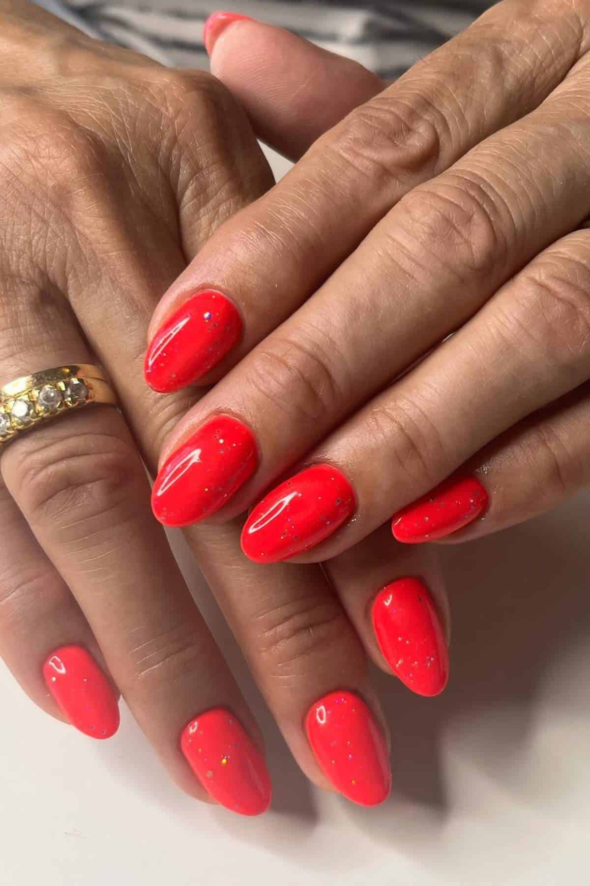 Hands with short, almond-shaped nails painted in glossy bright red polish with subtle glitter. One hand wears a gold ring with embedded clear stones.
