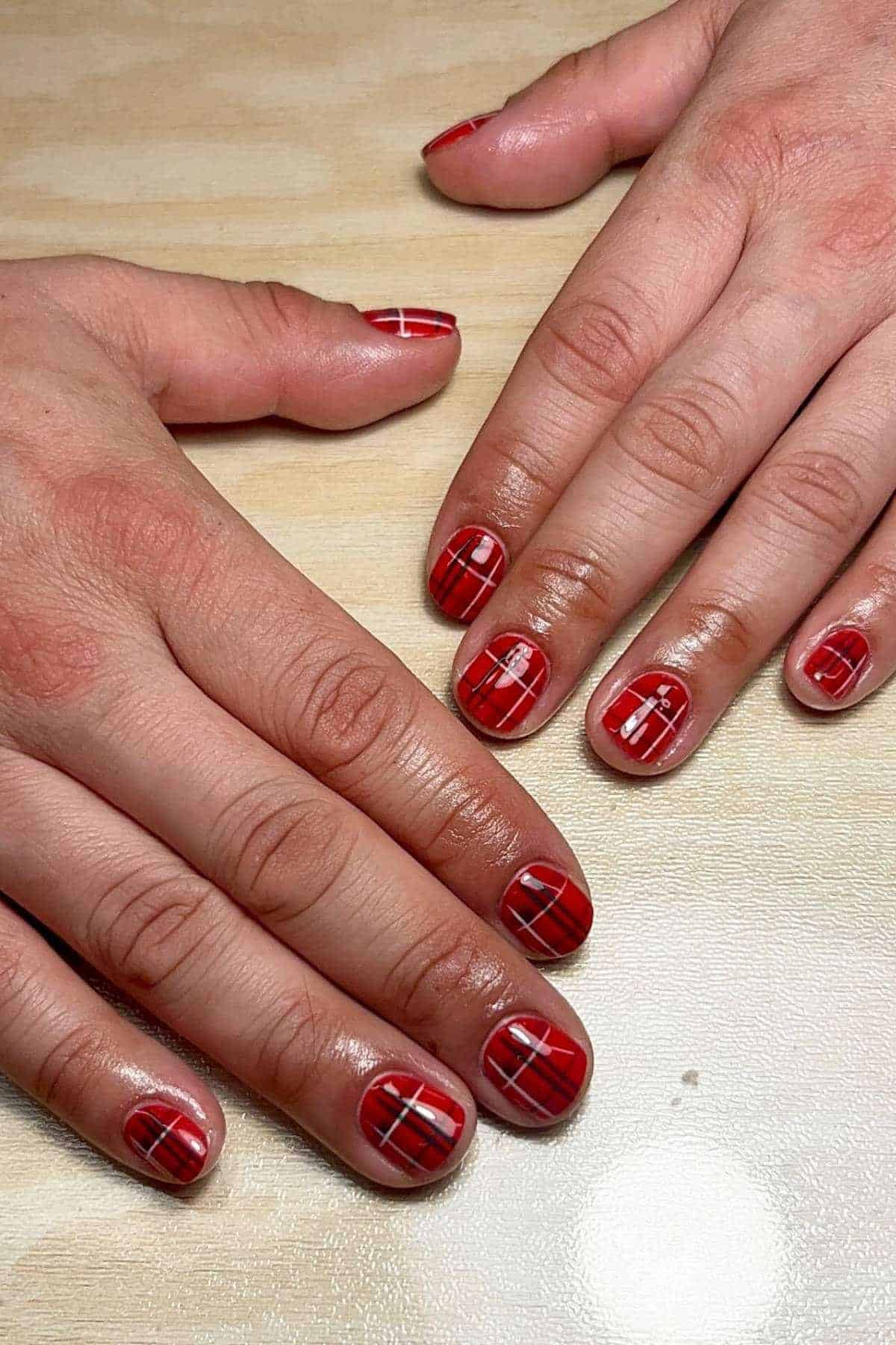 Hands with short nails painted red, featuring black and white plaid line designs, resting on a light wood surface.