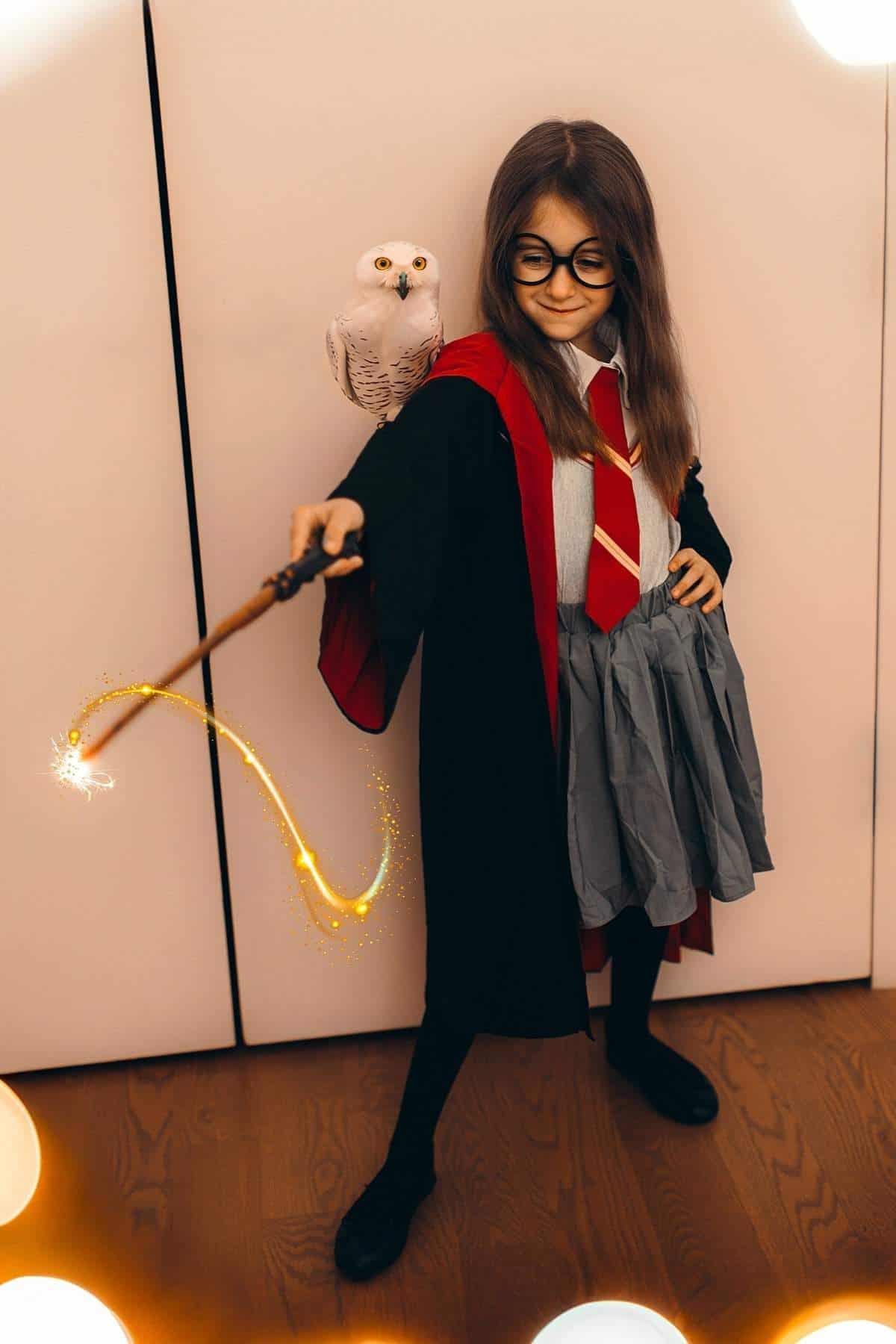 A child dressed as a wizard in glasses and a robe holds a wand with a glowing effect, with a toy owl perched on their shoulder.