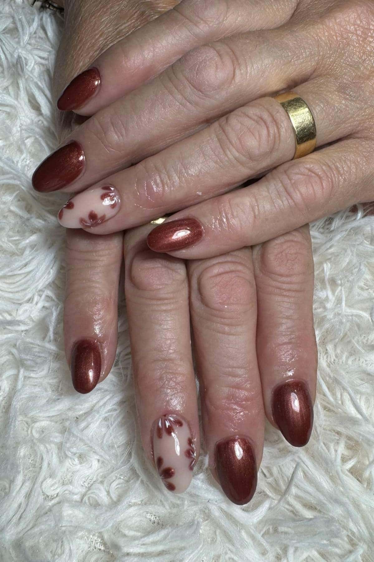 Hands with short, almond-shaped nails painted in metallic brown polish; two nails feature a white base with brown floral designs. The hands are resting on a white textured surface.