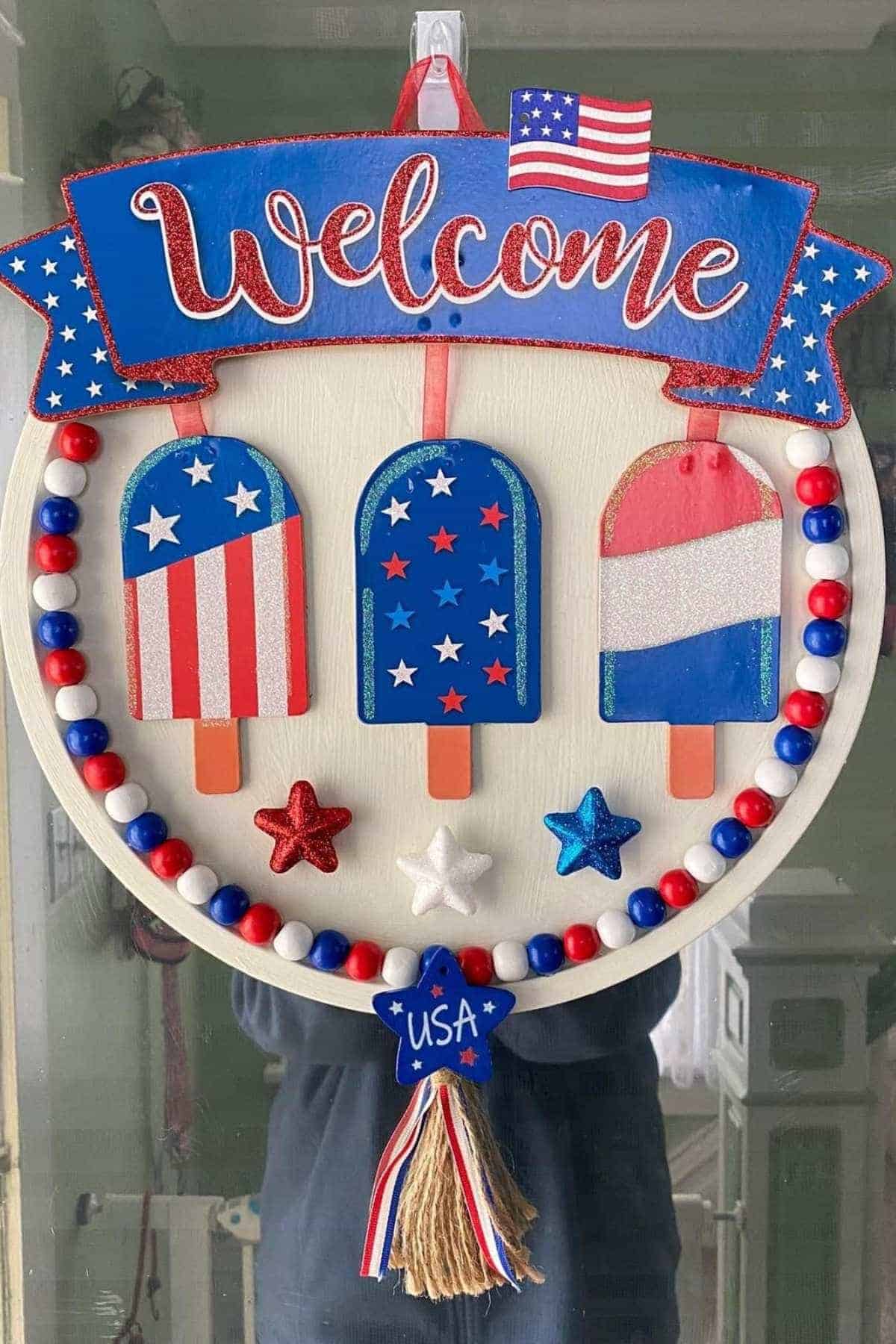 A door wreath with "Welcome" sign, three popsicle shapes in red, white, and blue, and USA-themed decorations, including stars and beads.