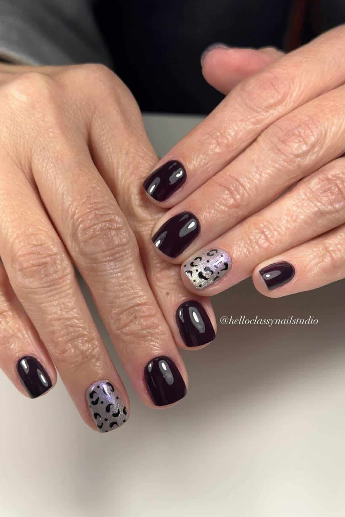 Hands with short, dark red manicured nails; one accent nail on each hand features a silver base with black leopard print design.