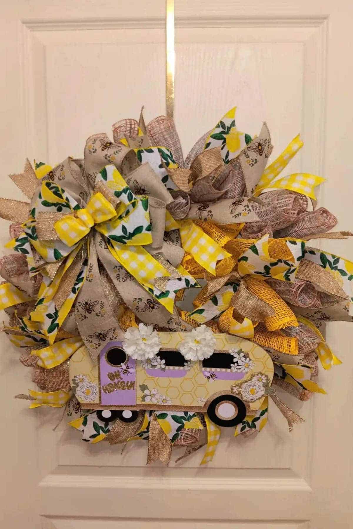 A decorative wreath with yellow, brown, and green ribbons features a cutout of a lavender truck with white flowers on a white door.