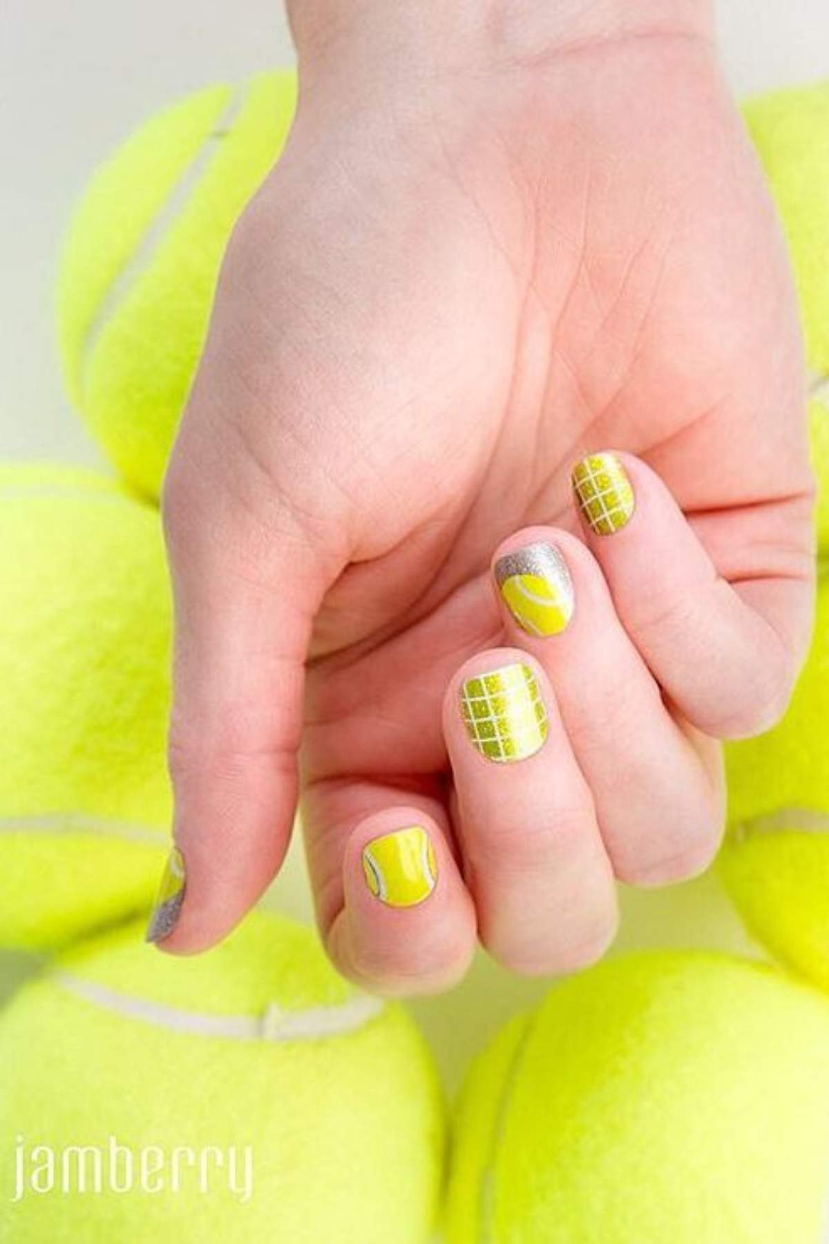 A hand with tennis-themed nail art is held against a background of stacked yellow tennis balls. The nails feature green, yellow, and white designs resembling tennis courts and balls.