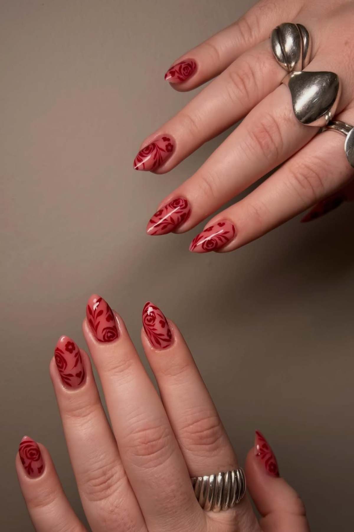 Close-up of hands with long, almond-shaped nails featuring red rose designs on a nude base; both hands are adorned with chunky silver rings.