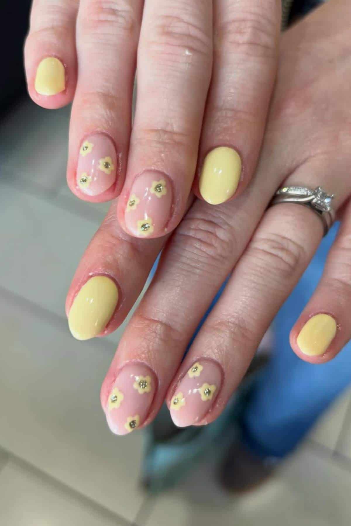 A hand with short, oval-shaped nails painted yellow and nude, featuring small yellow flower designs on some nails. A silver ring is on the ring finger.