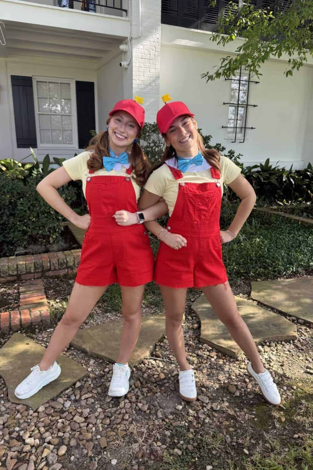 Two women stand outside in matching costumes with red overalls, yellow shirts, blue bow ties, red caps, and white shoes, posing with one leg forward and hands on hips.