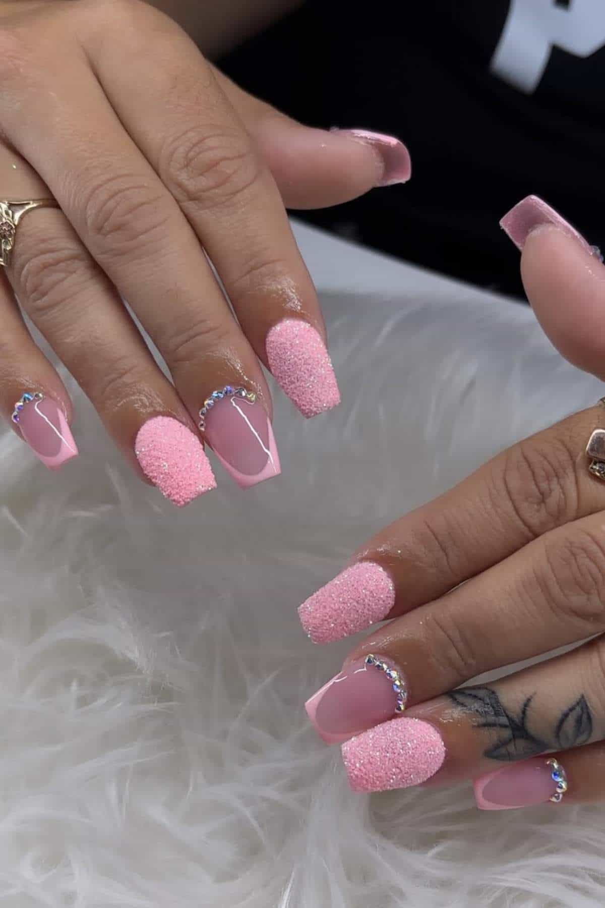 Hands with long square pink nails, some featuring textured glitter, others with smooth pink polish and rhinestone accents. Rings and a tattoo are visible on the fingers.