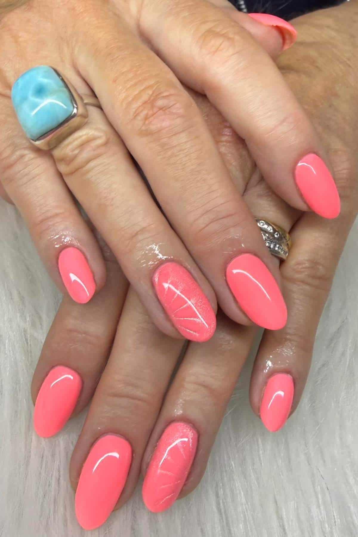 Hands with coral pink almond-shaped nails, one nail featuring a subtle leaf design. The person wears a silver ring with a blue stone and a gold ring with small diamonds.