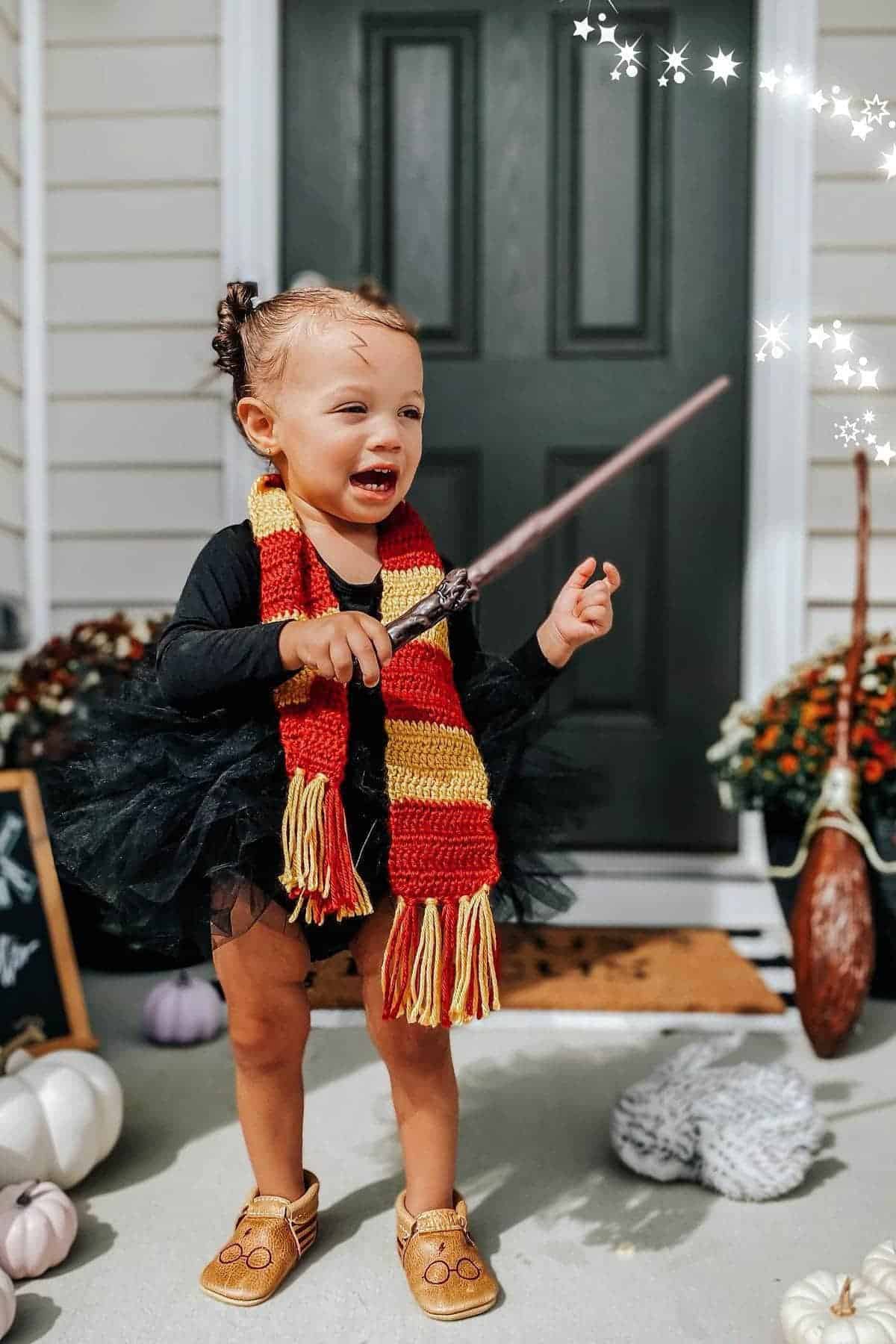 A young child wearing a black outfit and a red-and-yellow scarf holds a wand, standing in front of a door with autumn decorations and white pumpkins nearby.