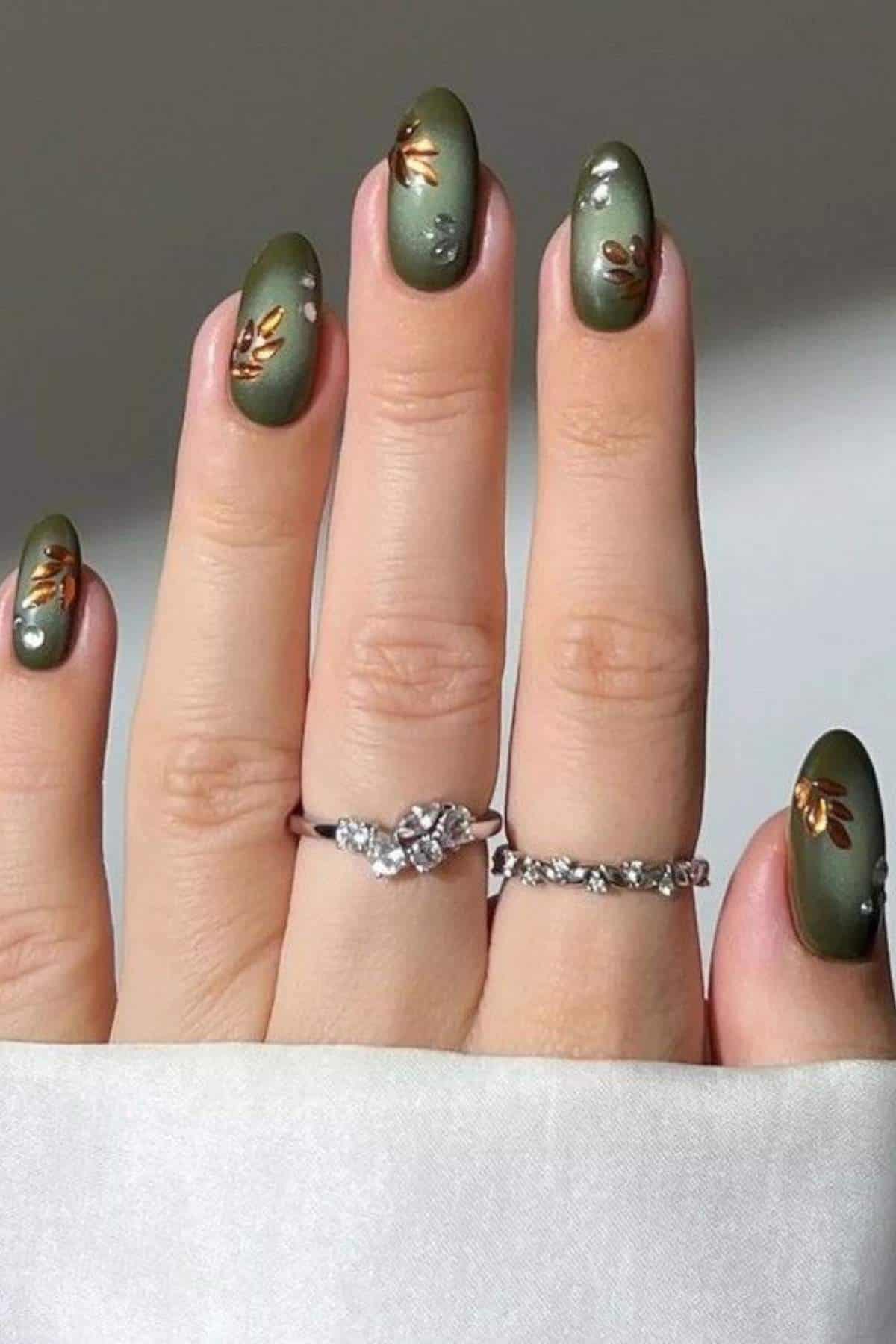 A hand with green matte almond-shaped nails featuring gold leaf and pearl accents, wearing two silver rings with gemstones.