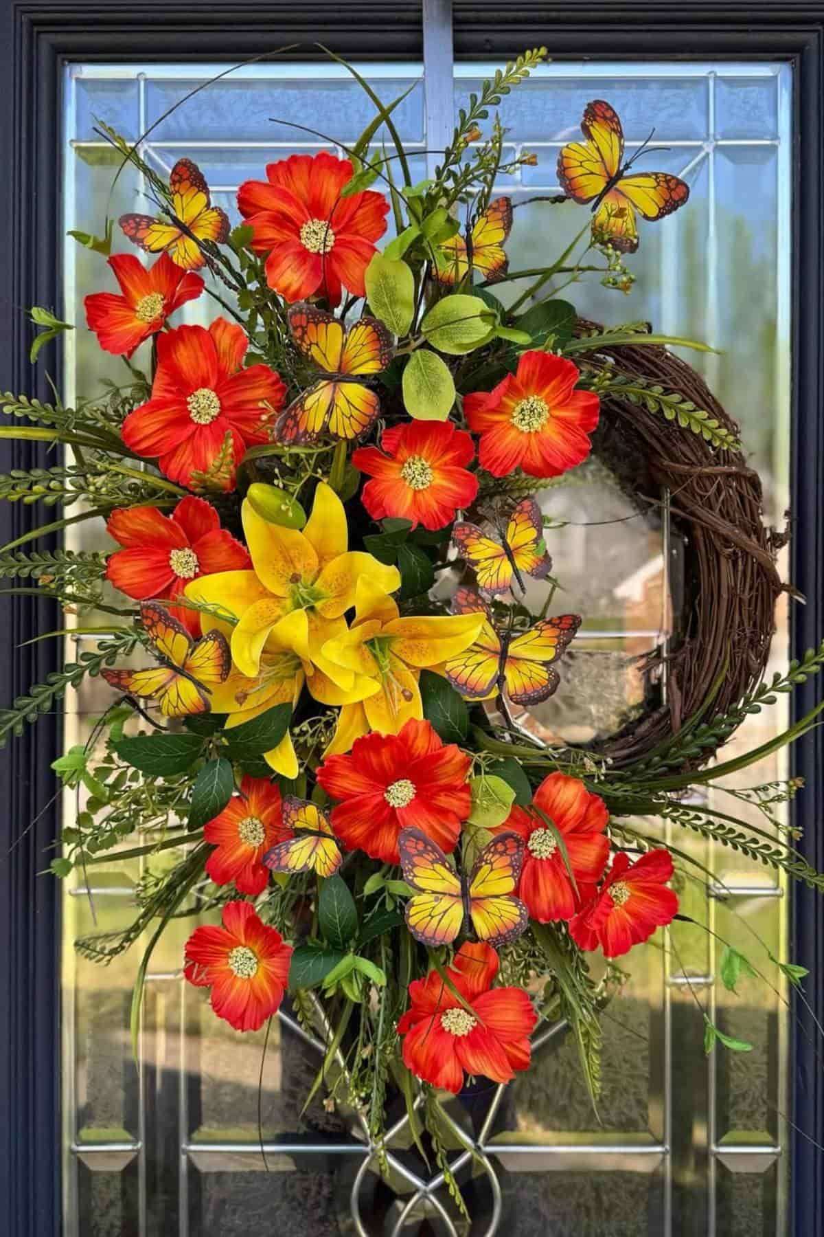 A floral wreath with bright red-orange flowers, a cluster of yellow lilies, greenery, and decorative butterfly accents hangs on a glass-paneled door.