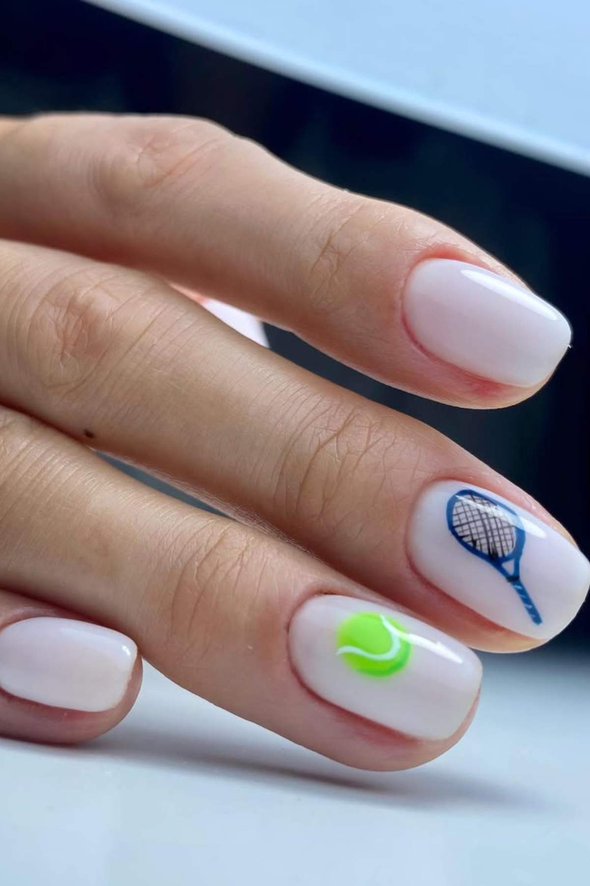 A hand with short, white-manicured nails; one nail features a small blue tennis racket and another has a green tennis ball design.