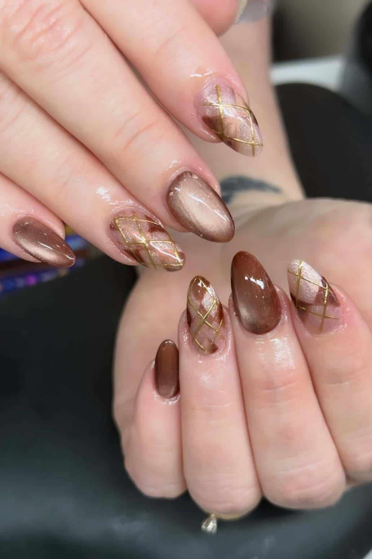 Hands with almond-shaped nails featuring brown polish and accent nails with gold crisscross lines over a shimmery base.