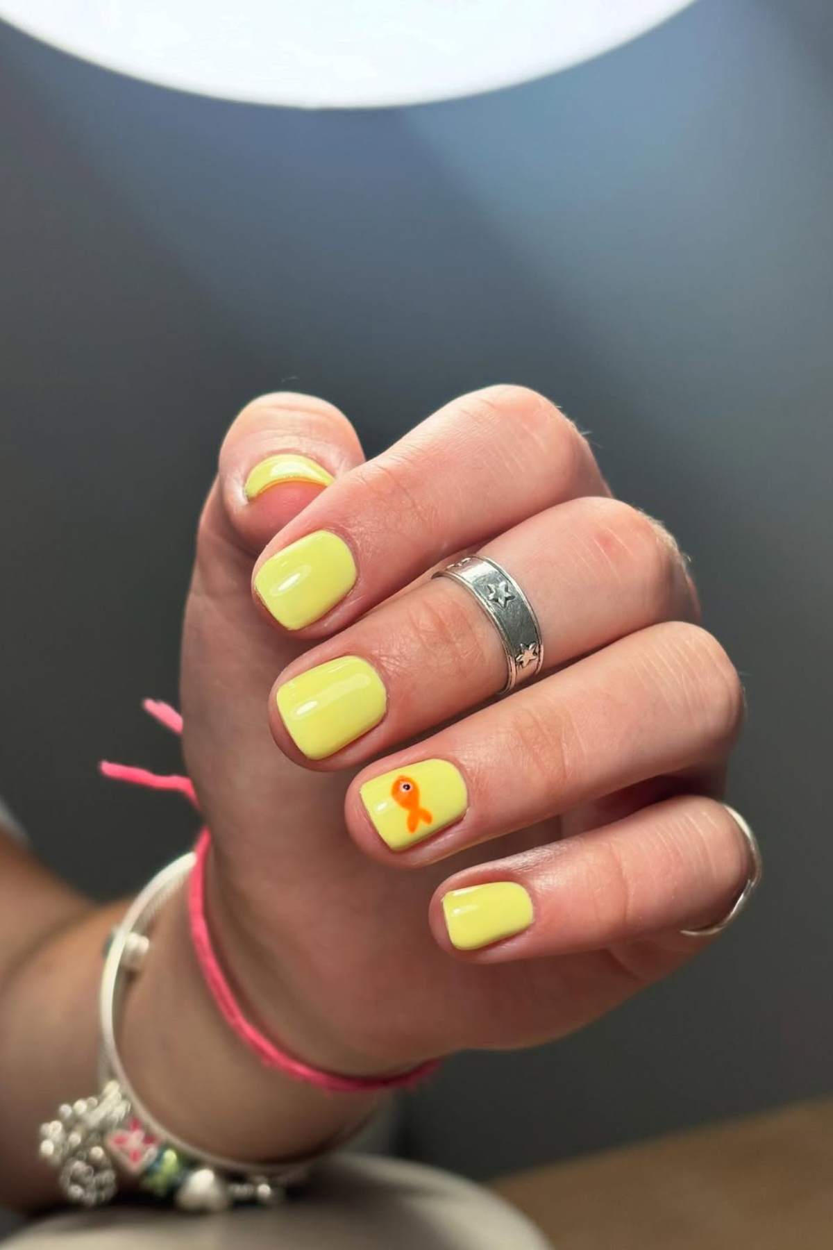 A hand with short, square-shaped nails painted yellow; one nail features a small orange fish design. The person wears rings and bracelets.