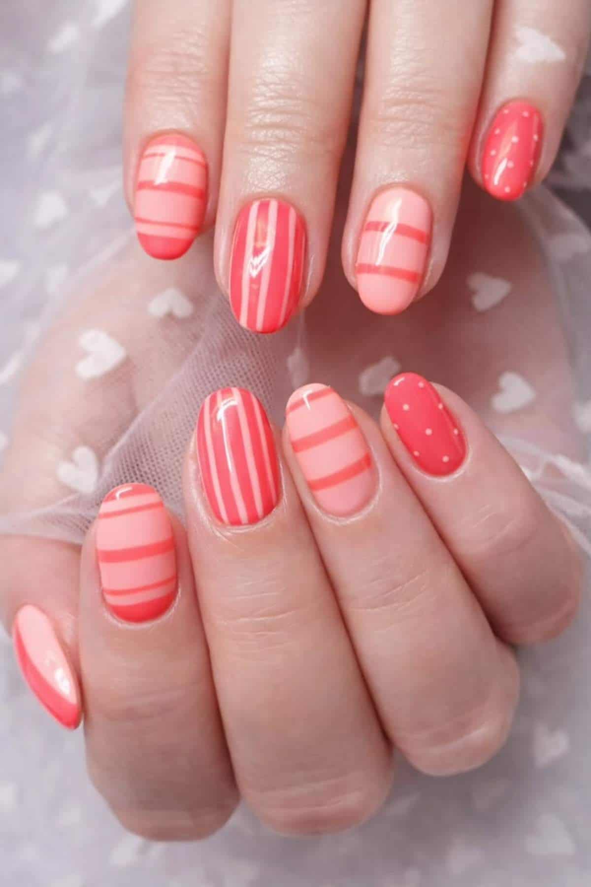 Hands with oval-shaped nails painted in shades of pink and coral, featuring a mix of stripes and polka dot nail art designs.