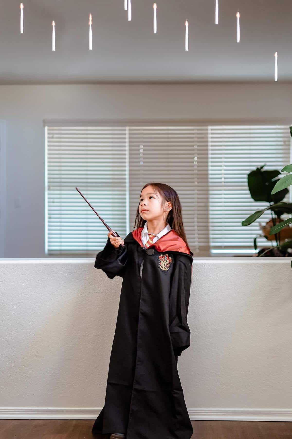 Young girl in a Hogwarts robe and tie holds a wand, looking up at floating candles.
