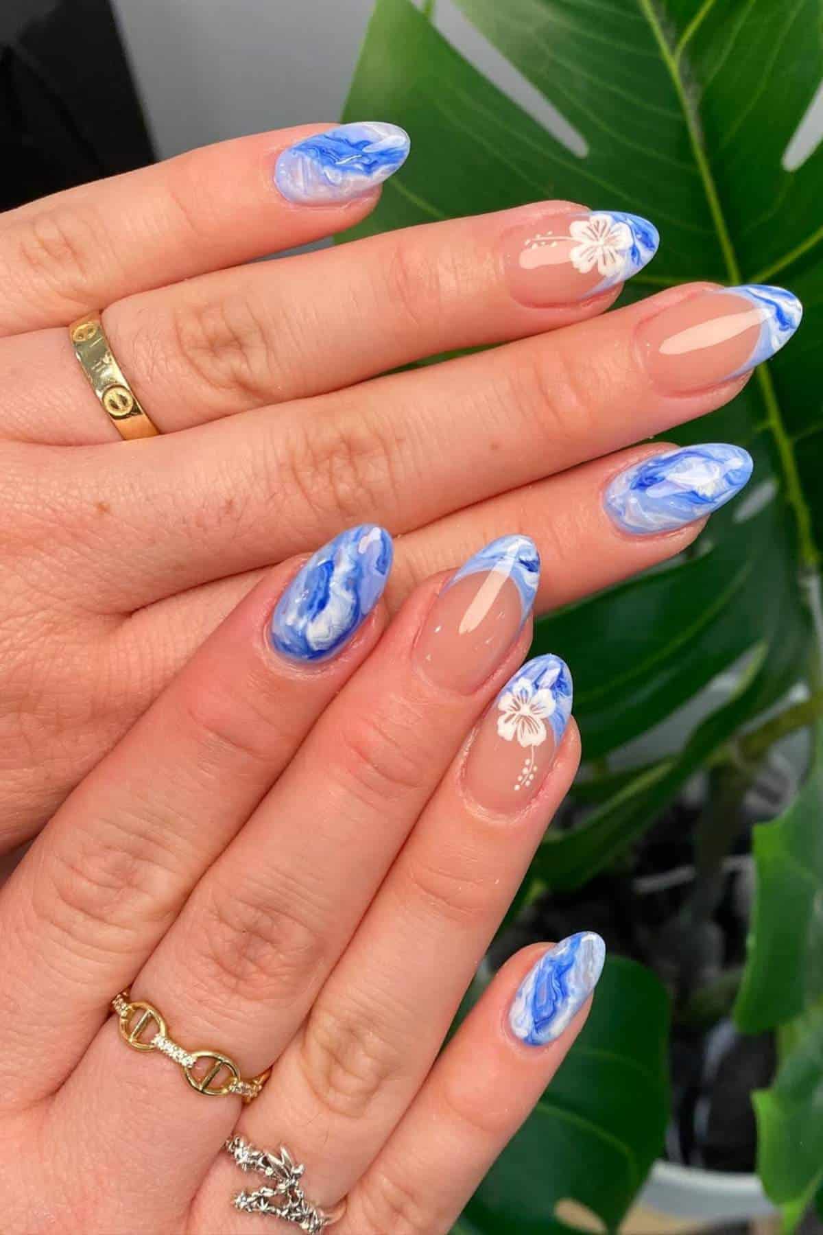 Hands with long almond-shaped nails featuring blue and white marble designs and small white flower accents. Green leaf is visible in the background.