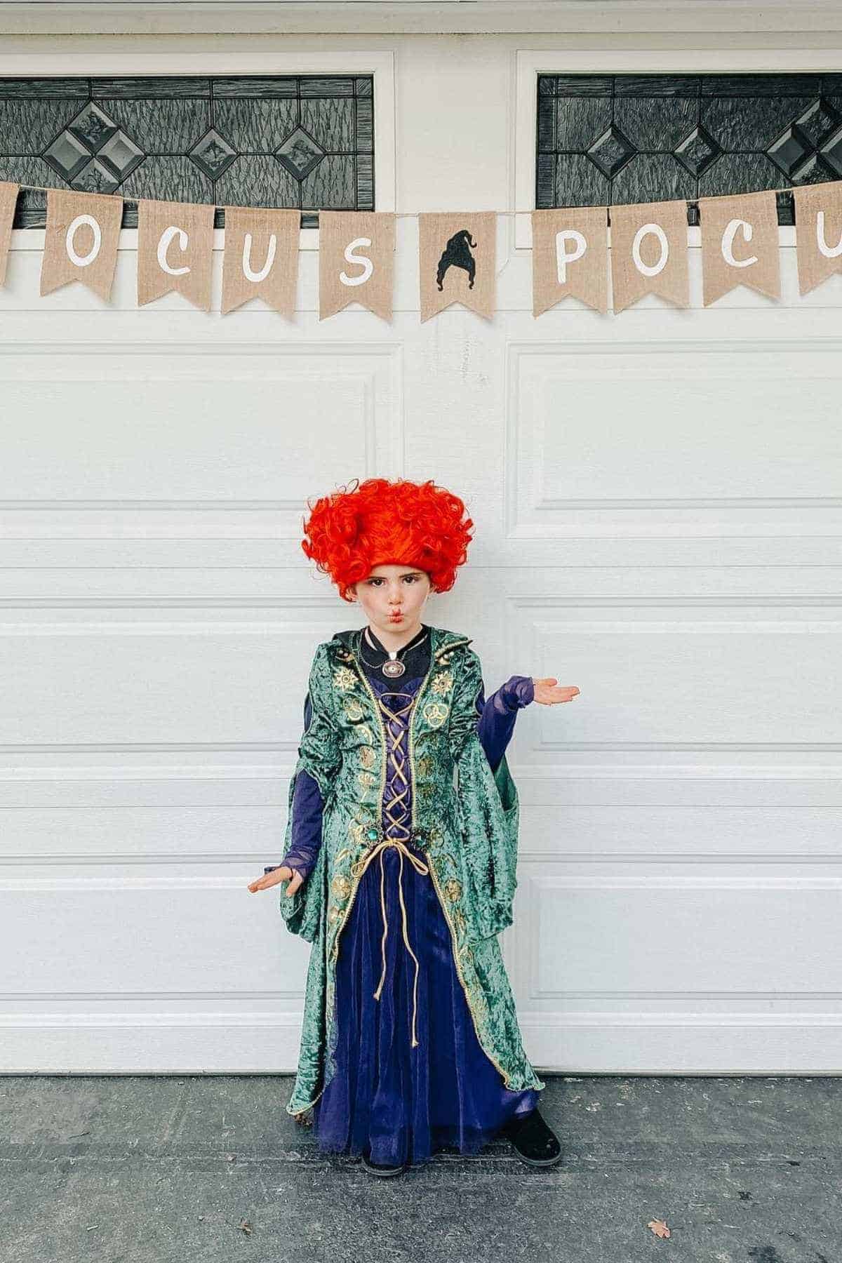 Child in a green and purple witch costume with a large red wig poses in front of a garage decorated with a "Hocus Pocus" banner.