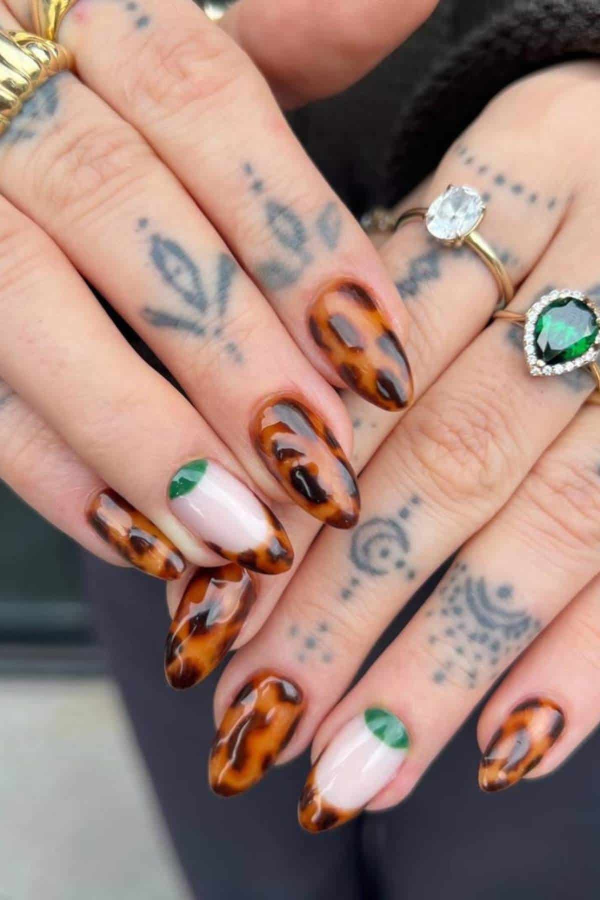 Close-up of hands with almond-shaped nails featuring a tortoiseshell design and green crescent accents, alongside tattooed fingers and multiple rings.