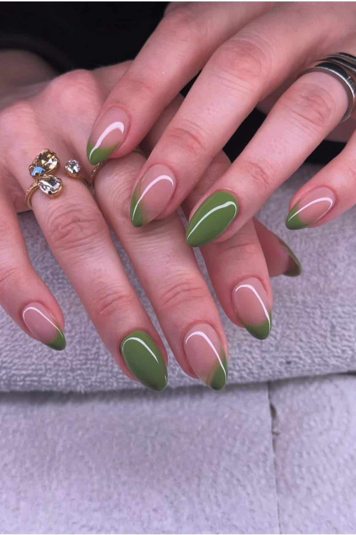 Hands with almond-shaped nails featuring a green French tip manicure, resting on a light textured surface. The wearer has rings on their fingers.