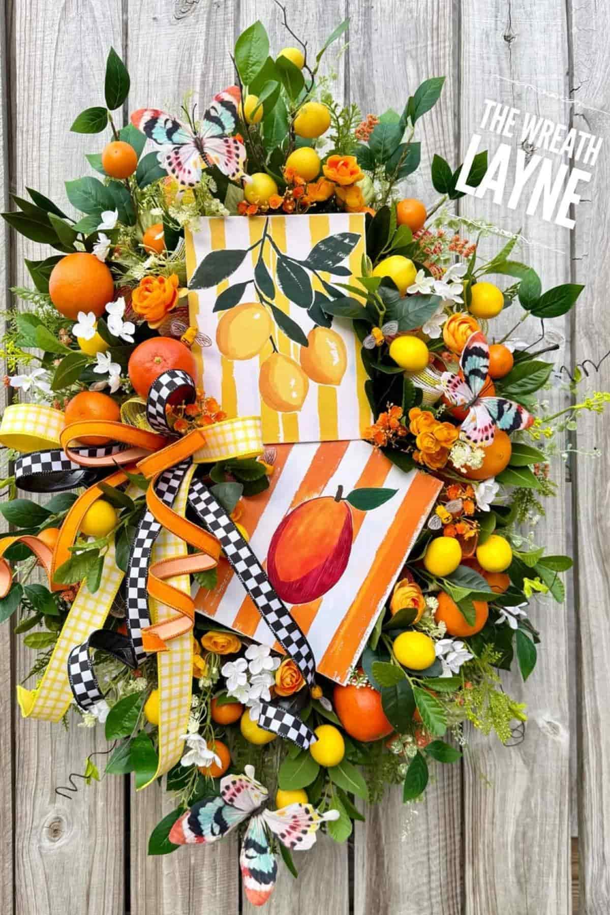 A decorative wreath with artificial lemons, oranges, flowers, ribbon bows, butterfly accents, and citrus-themed signs mounted on a wooden background.