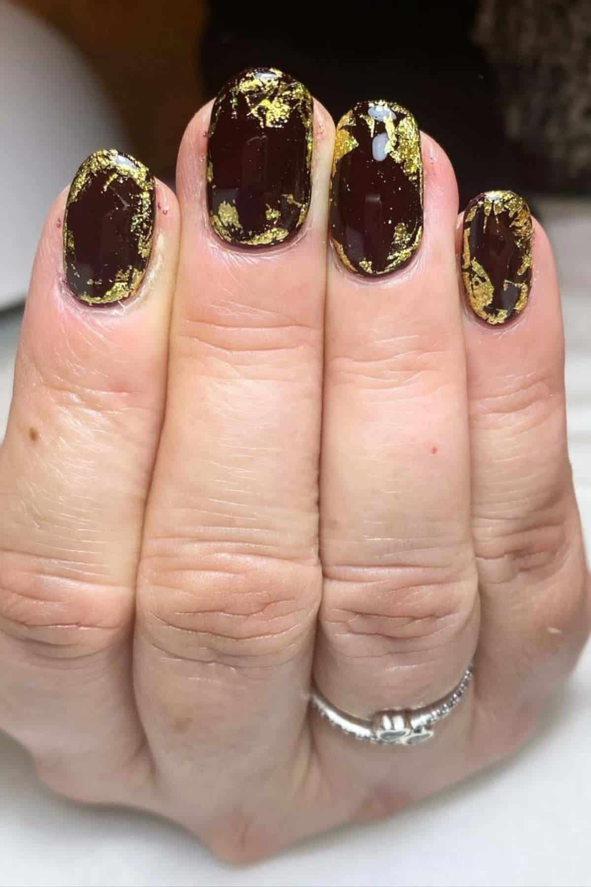 A hand with short, rounded nails painted dark brown and decorated with irregular gold foil accents.