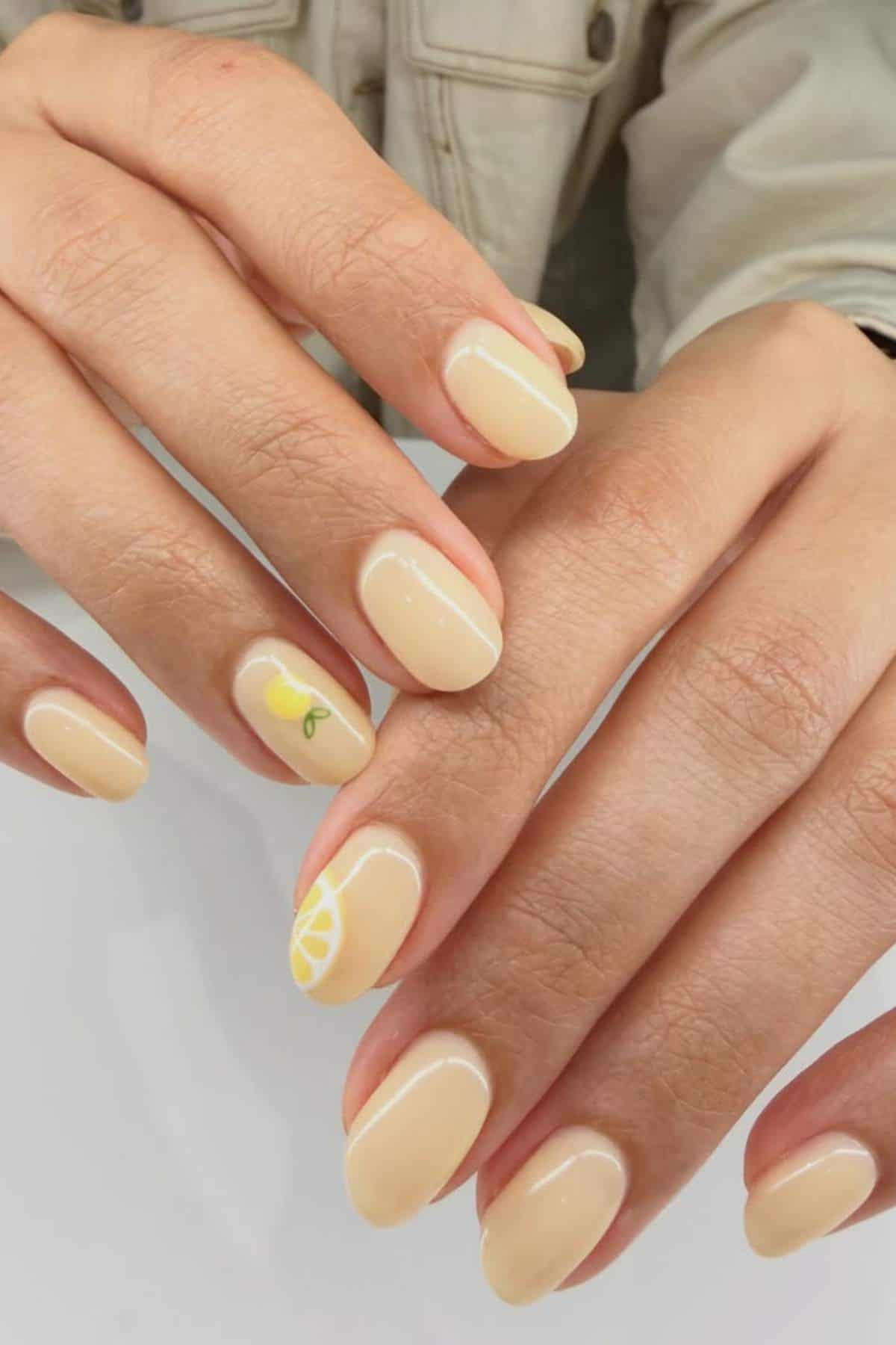 Hands with short, oval-shaped nails painted in pale yellow polish; one nail has a small lemon slice design, another has a small lemon with leaf detail.