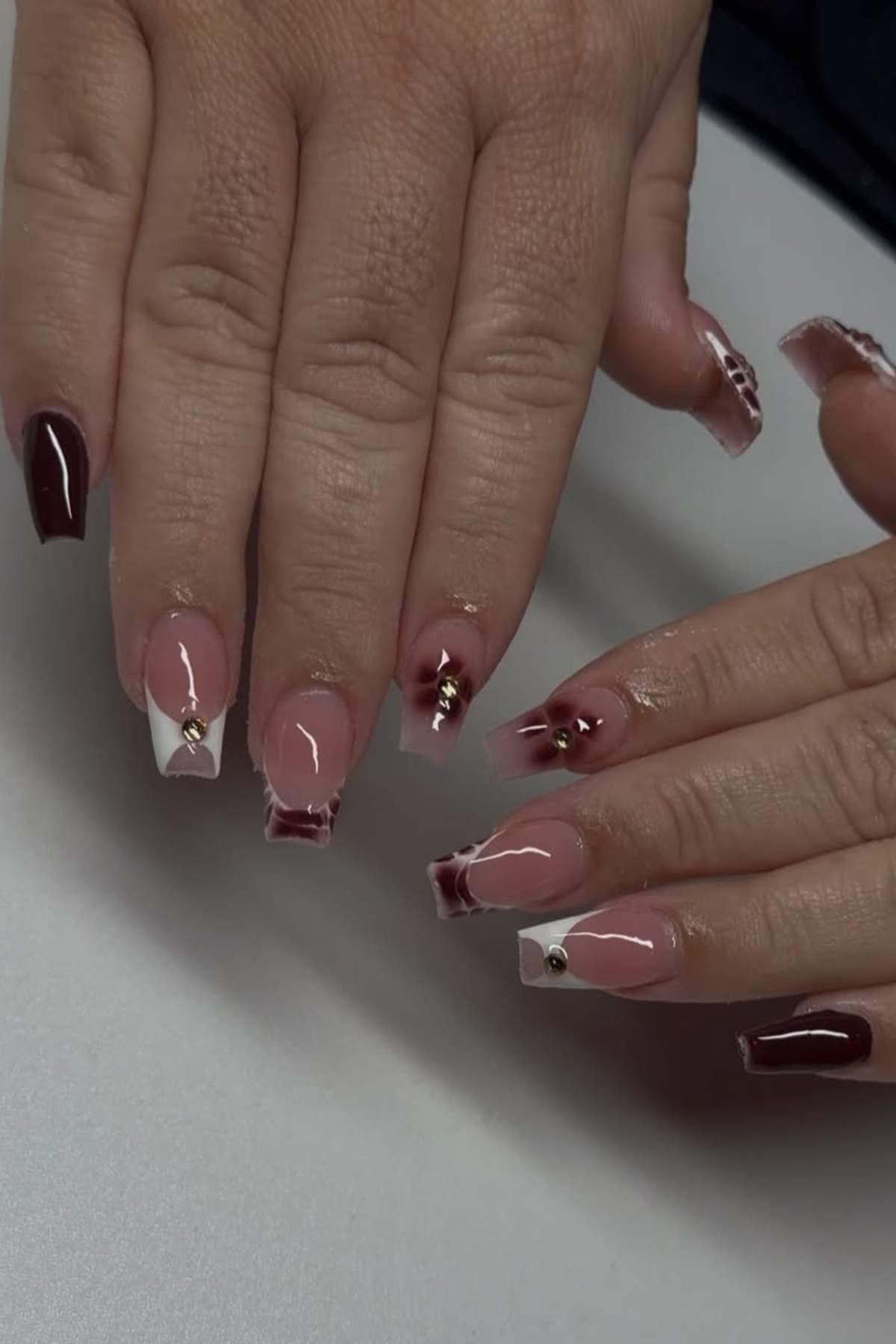 Hands with long acrylic nails featuring burgundy, nude, and white geometric designs, gold studs, and glossy finish, displayed on a white surface.