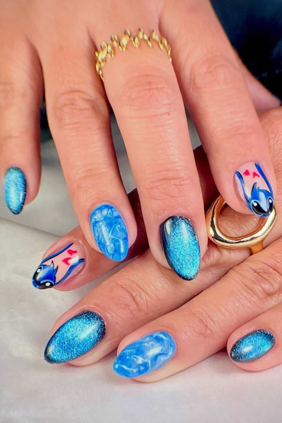 Close-up of hands with blue manicured nails featuring metallic blue polish, marble patterns, and koi fish designs; person wears gold rings.