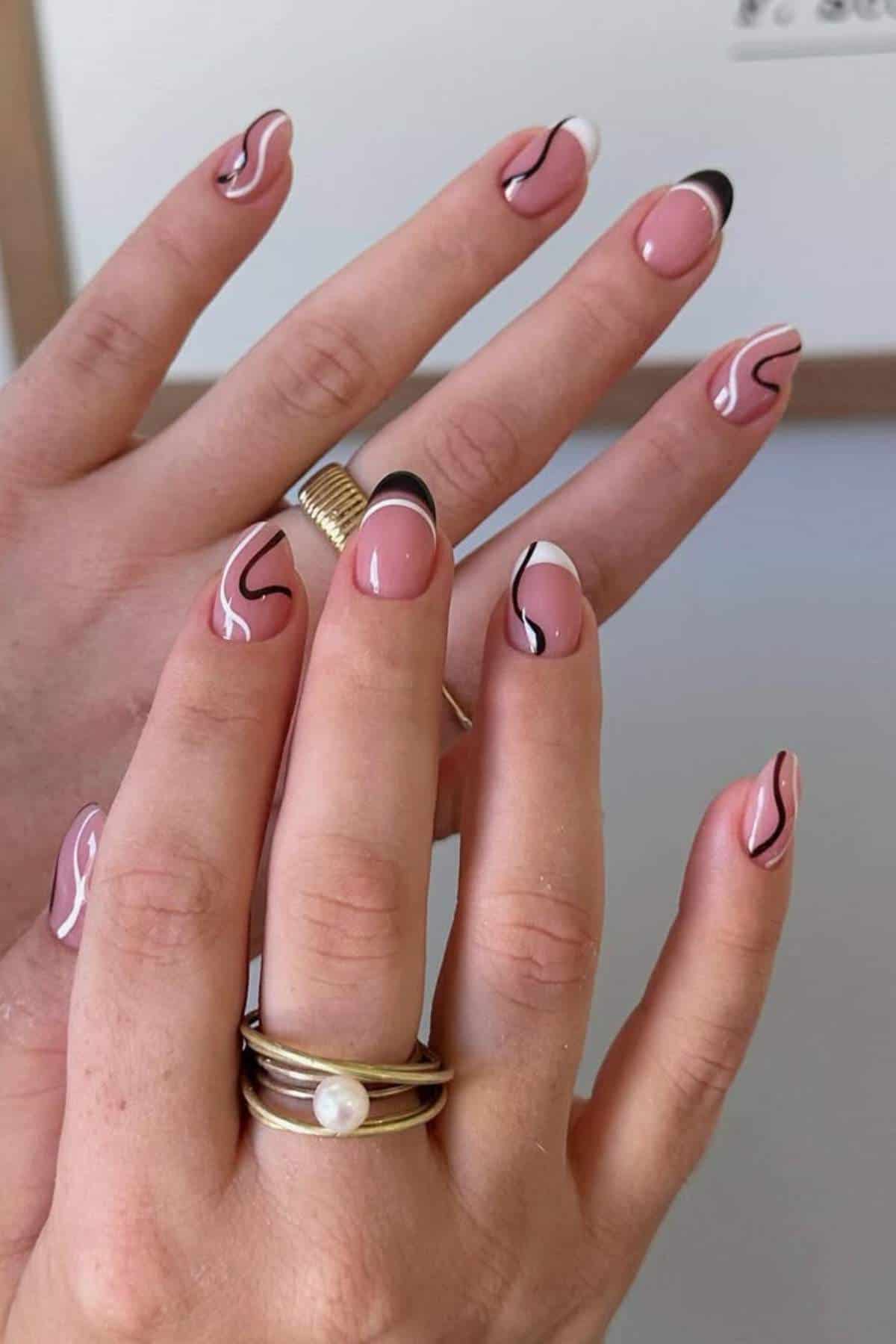 A hand with nude pink nails featuring black and white abstract wavy line designs and french tips, wearing multiple gold rings.