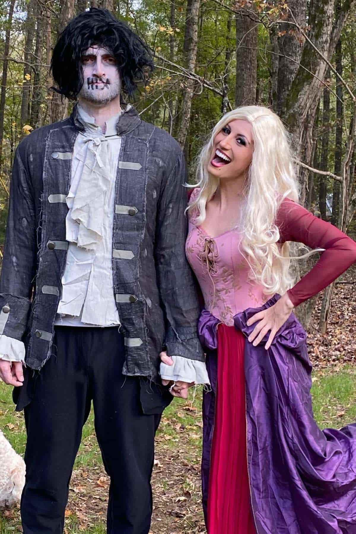 Two people in costume stand outdoors; one dressed as a zombie with pale makeup and dark clothes, the other as a princess with a long blonde wig and a purple and pink gown.
