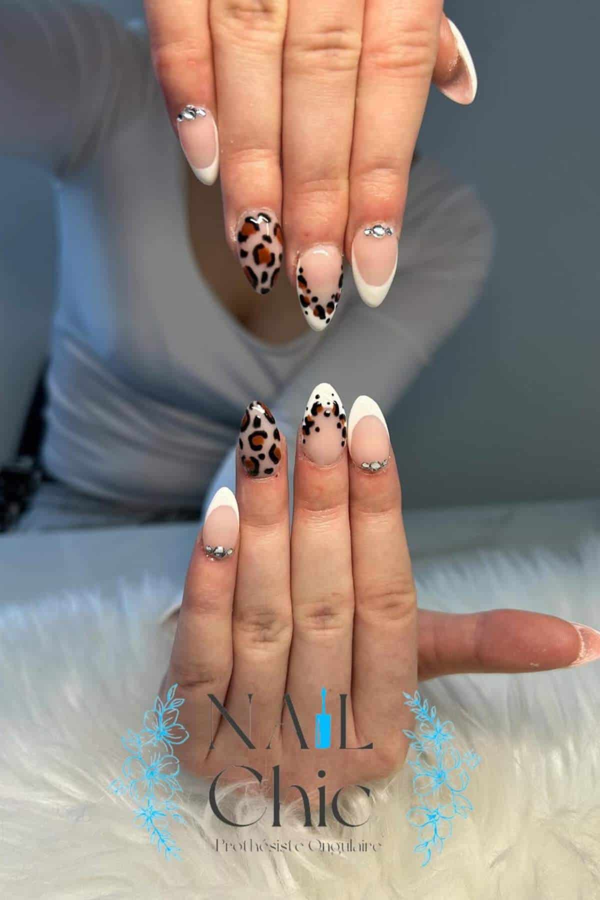 A person displays manicured hands with almond-shaped nails featuring leopard print accents, nude polish, white tips, and rhinestone decorations.