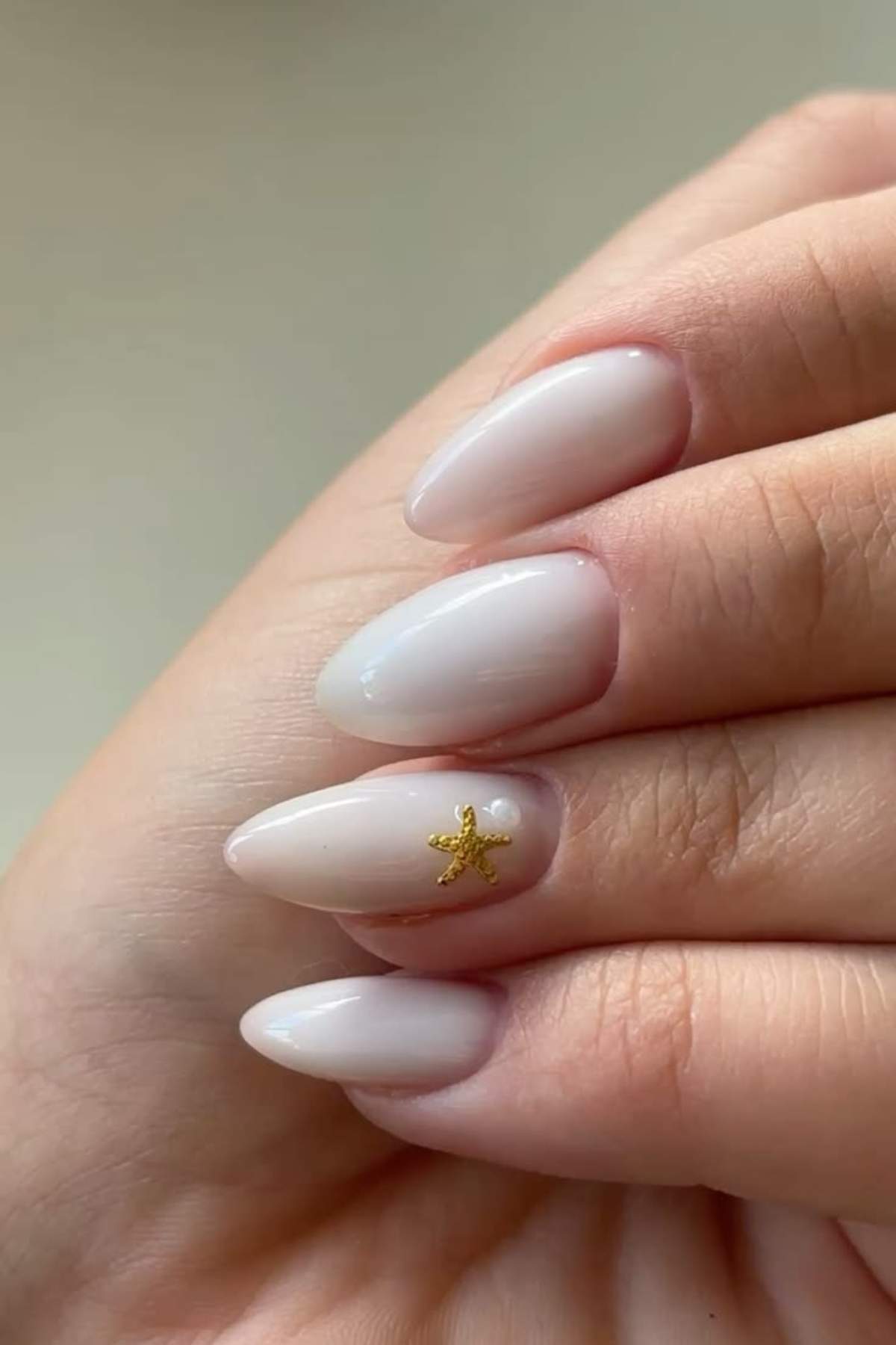A hand with almond-shaped nails painted in a pale nude color, featuring a small gold starfish embellishment on the ring fingernail.