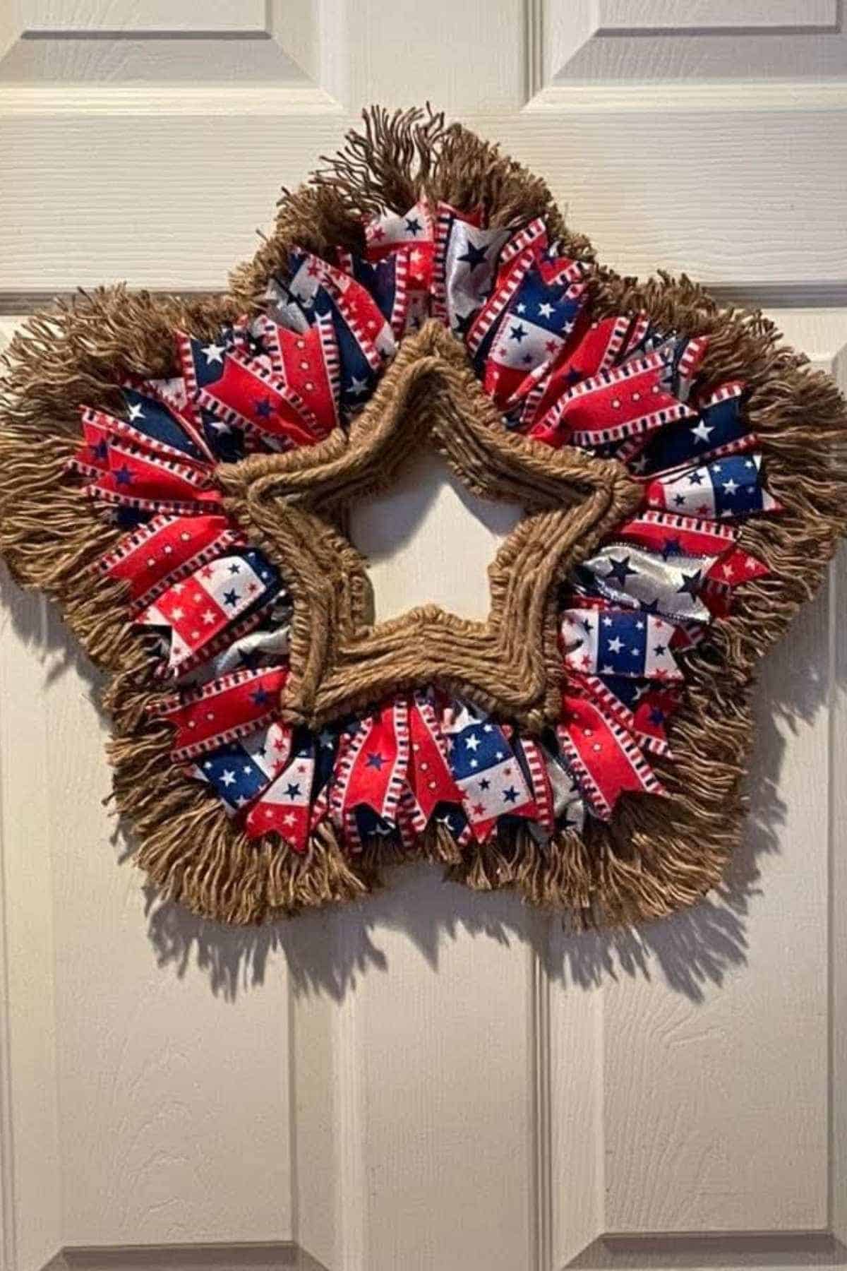 A star-shaped wreath made of rope and patriotic red, white, and blue ribbon, hanging on a beige door.